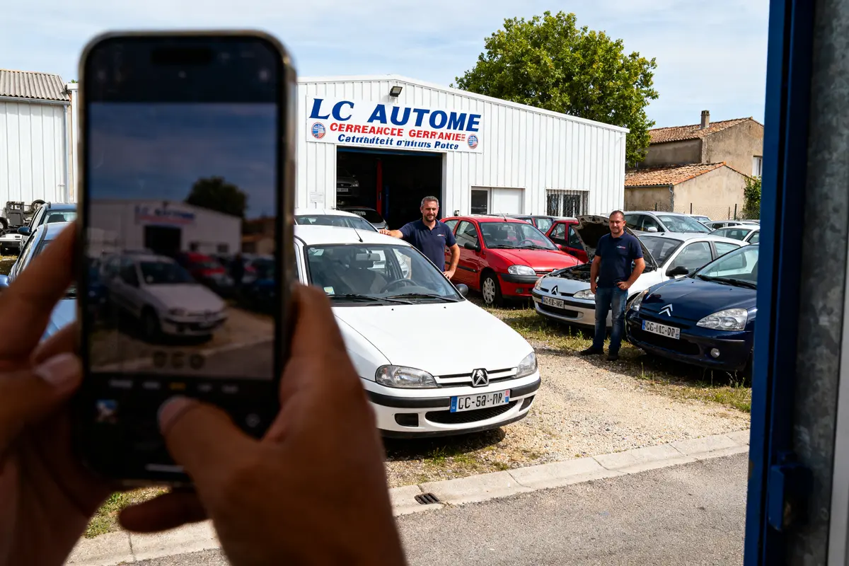 Deux hommes devant garage automobile, plusieurs voitures garées autour. Photo prise sur smartphone tenant l'image large.