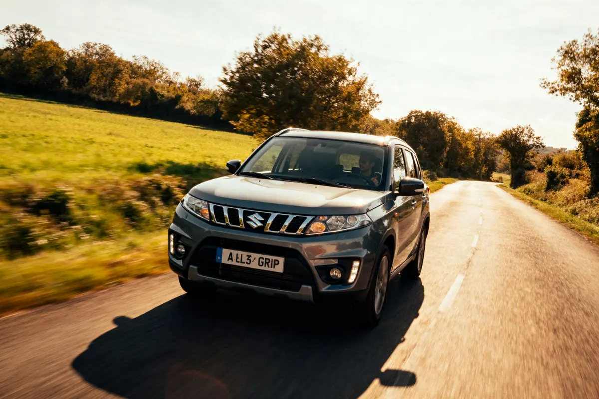 SUV Suzuki Vitara gris roulant sur route de campagne ensoleillée, entourée de verdure et arbres.