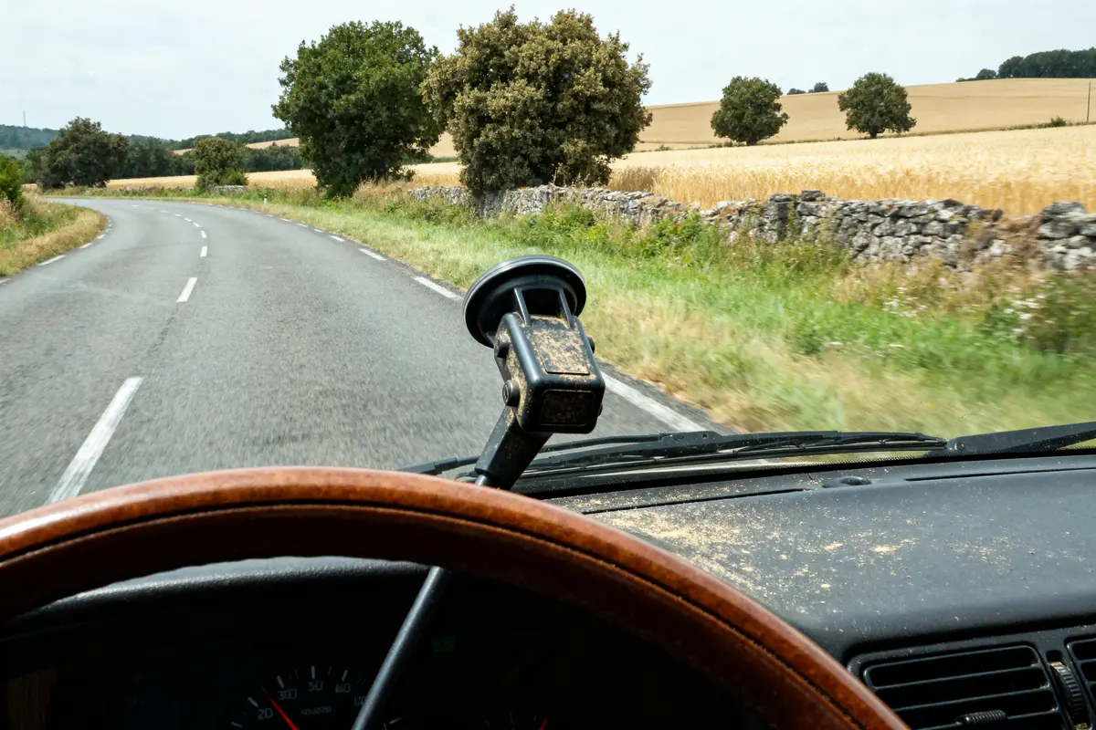 Vue d'une route de campagne depuis une voiture, volant en premier plan, champs et arbres alentours.