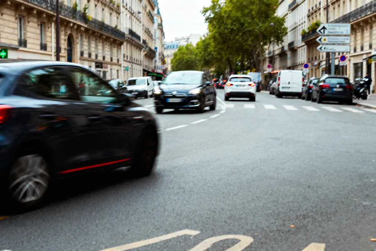 Rue urbaine mouvementée avec voitures en circulation, bâtiments haussmanniens, arbres en arrière-plan. Jour.