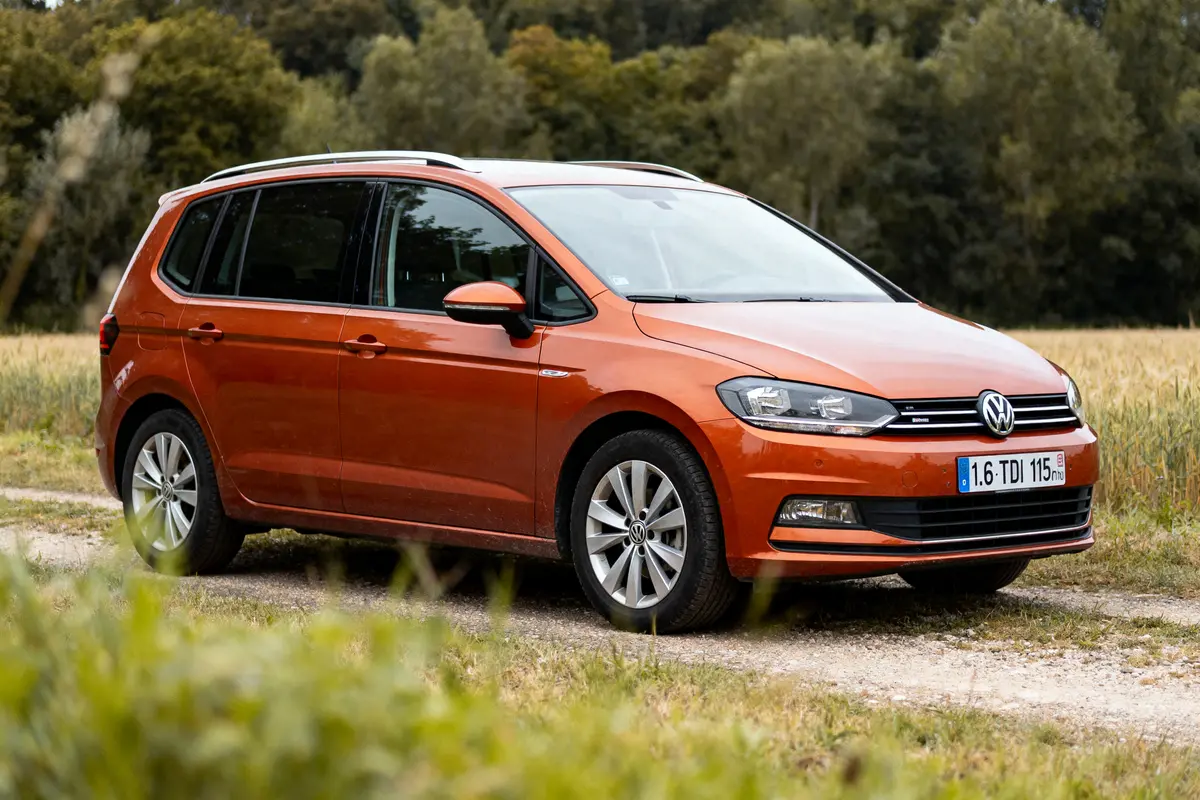 Voiture orange Volkswagen sur chemin de campagne, entourée de verdure et forêt en arrière-plan.