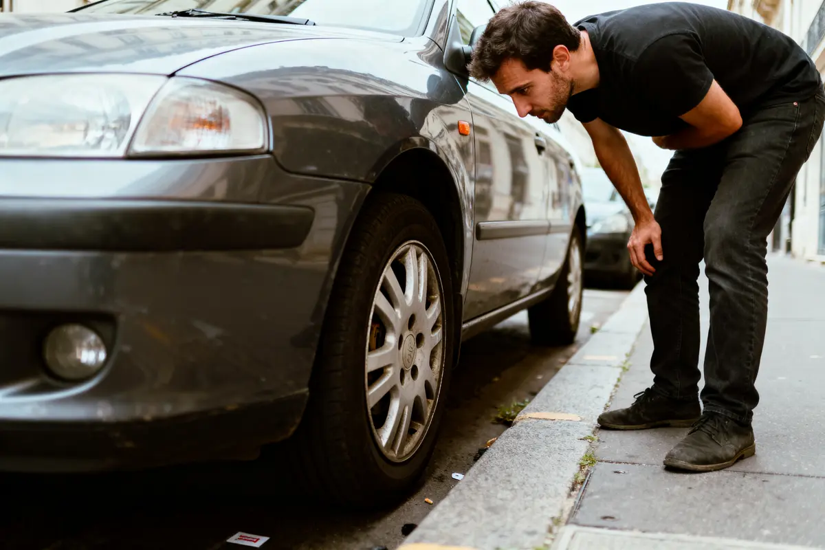 Homme en T-shirt noir examinant roue avant d'une voiture grise garée sur trottoir urbain.
