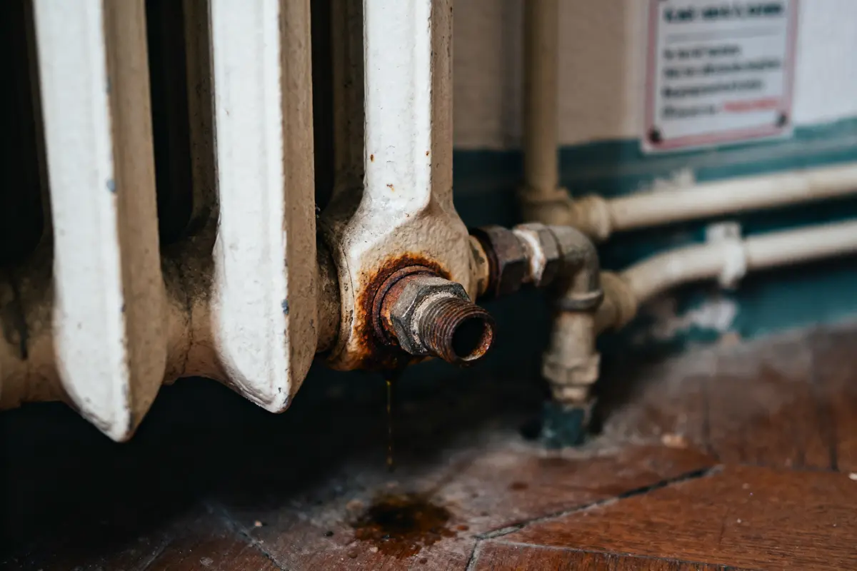 Radiateur en métal avec tuyau rouillé et fuite d'eau, formant une flaque sur un sol en bois.