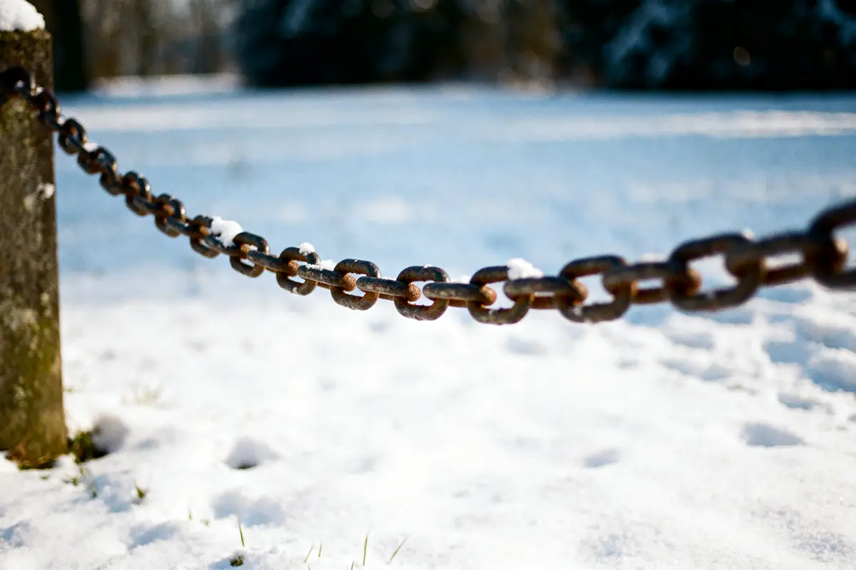 Chaîne rouillée traversant un paysage enneigé, attachée à un poteau. La neige recouvre légèrement la chaîne.