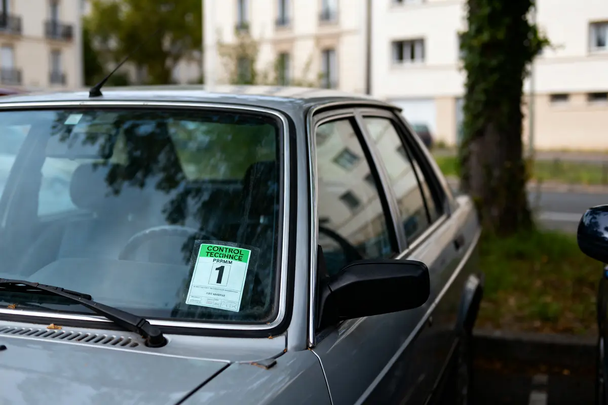 Voiture grise avec vignette de contrôle technique sur pare-brise, stationnée près de bâtiments résidentiels.