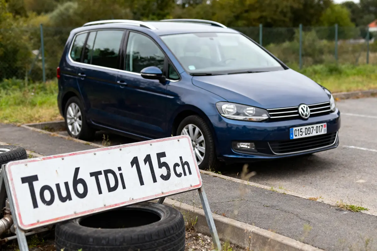 Voiture bleue Volkswagen garée près d'un panneau "Tou6 TDI 115ch" et des pneus.