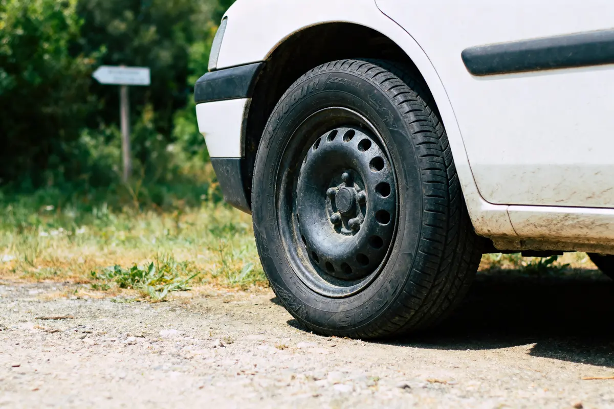 Roue avant droite d'une voiture blanche sur chemin de terre, panneau flou en arrière-plan.