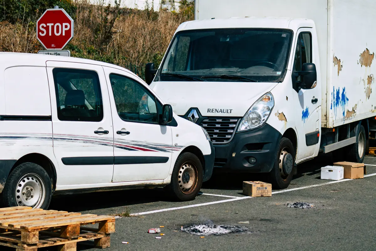 Deux camionnettes blanches garées, panneaux abîmés, palettes en bois, boîte, mégots, panneau "STOP" en arrière-plan.