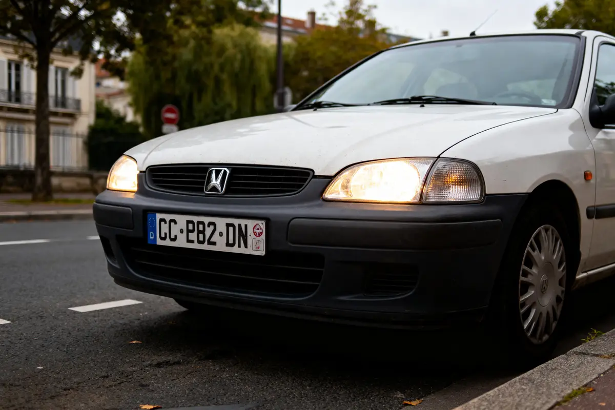 Voiture blanche Honda stationnée, phares allumés, rue bordée d'arbres et bâtiments résidentiels.