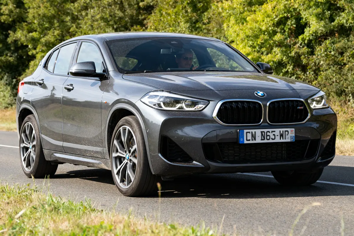 BMW X2 car gris roulant sur route bordée d'arbres, vue de devant